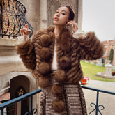 A woman wearing a brown fur coat and a patterned skirt stands on a balcony, with a stone building and a fountain visible in the background.