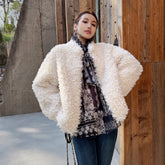 A woman wearing a white fluffy coat and a patterned shirt stands in front of a wooden wall.