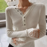 A person wearing a white sweater with lace detailing and buttons, standing in front of a window.