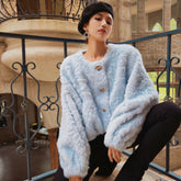 A woman wearing a light blue fur coat and black pants is posing on a balcony with a wrought iron railing in the background.