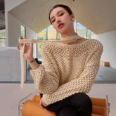 A woman wearing a beige sweater and holding a scarf around her neck is sitting on an orange chair in a room with large windows.