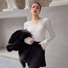 A woman wearing a white sweater with lace sleeves and a black fur coat stands in front of a white wall.