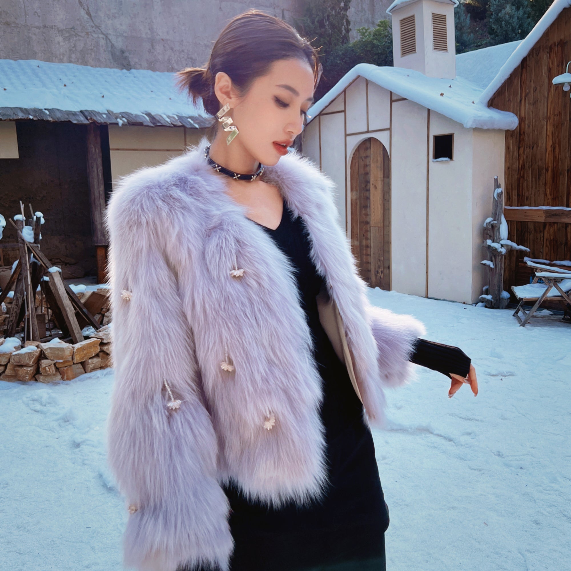 A woman wearing a fur coat and black dress stands in front of a snowy building.