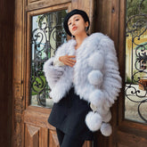 A woman wearing a white fur coat and black skirt stands in front of a wooden door with an ornate wrought iron design.