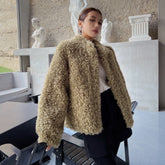 A woman wearing a beige furry coat stands in a room with a stone wall and statues in the background.