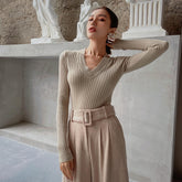 A woman wearing a beige sweater and a beige skirt stands in front of a white marble wall, with a statue visible behind her.