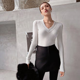 A woman wearing a white sweater and black skirt stands in front of a white wall, holding a black fur coat.