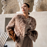 A woman wearing a fur coat and holding a brown purse stands in front of a wall with statues.