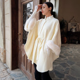 A woman wearing a white fur-trimmed poncho stands in front of a wooden door, with a potted plant visible in the background.