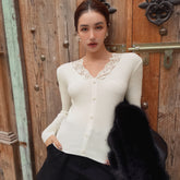 A woman wearing a white cardigan with lace trim and holding a black fur coat stands in front of a wooden door.