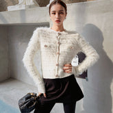 A woman wearing a white furry cardigan and black skirt, posing in front of a concrete wall. She is holding a black purse in her hand.