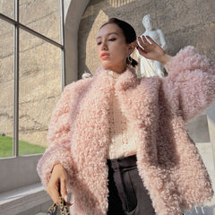 A woman wearing a pink fluffy coat stands in front of a window, with a statue visible in the background.