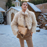 A woman wearing a fur coat and holding a beige handbag stands in front of a snowy landscape with a small wooden structure.