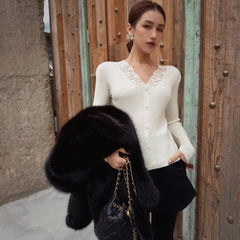 A woman wearing a white cardigan and holding a black fur coat stands in front of a wooden wall.
