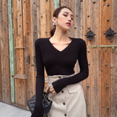 A woman wearing a black sweater and a beige skirt stands in front of a wooden door, holding a black handbag.