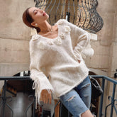 A woman wearing a white sweater with flower designs and ripped jeans is posing with her eyes closed and leaning against a wrought iron railing.