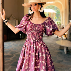 Woman in a purple floral dress and hat standing indoors.
