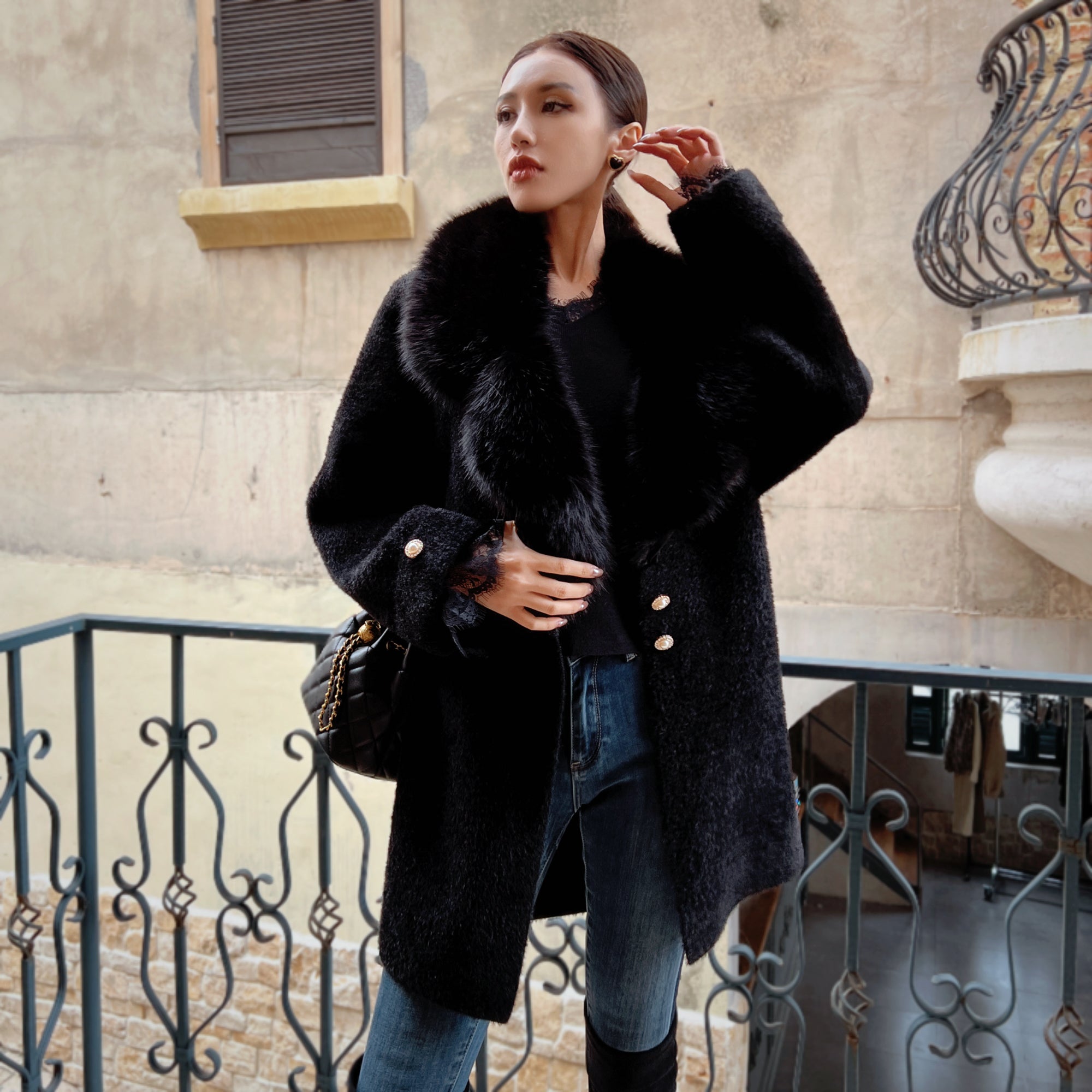 A woman wearing a black fur coat stands on a balcony, looking off to the side.
