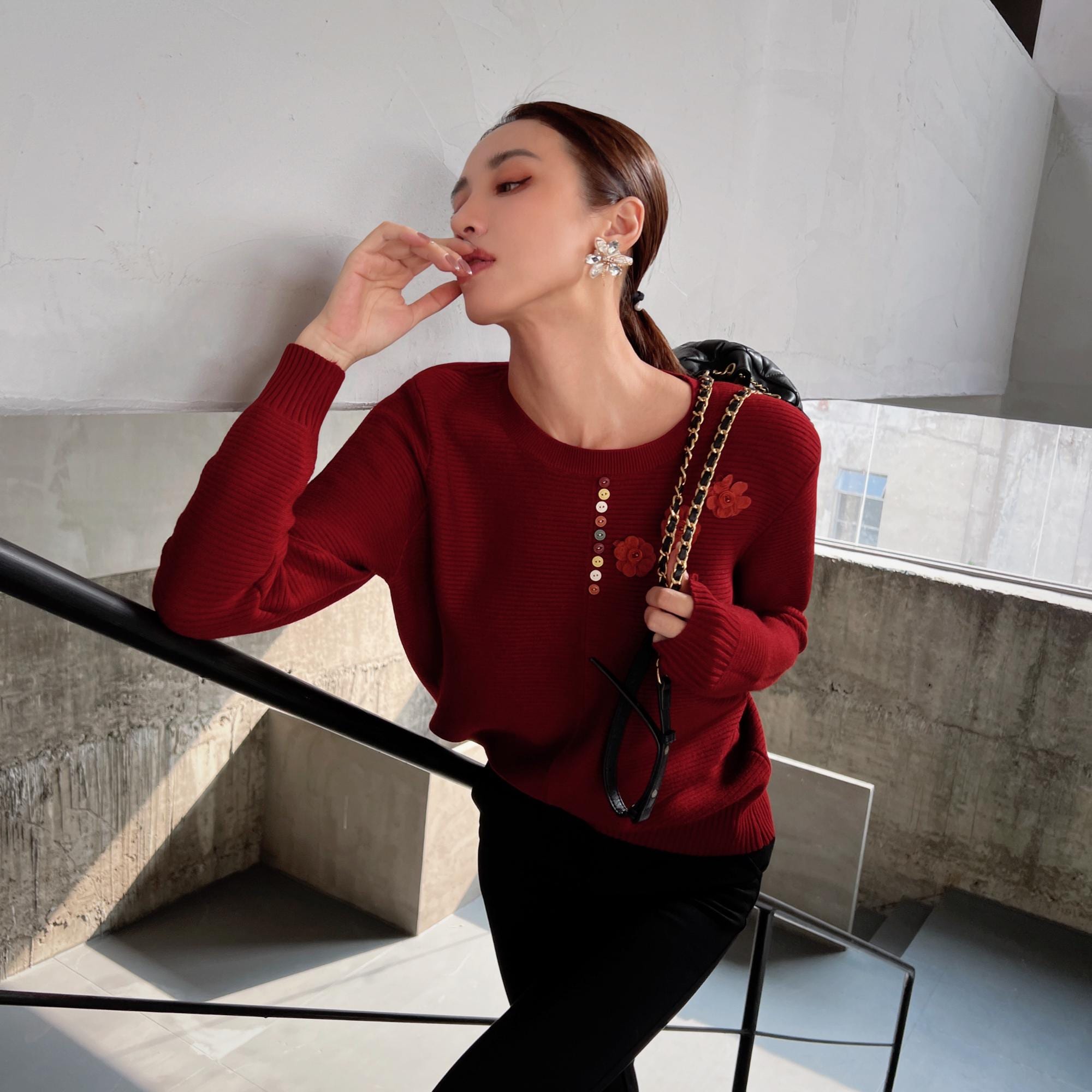 A woman in a red sweater stands on a staircase, looking down at her hand, with a black purse hanging from her shoulder.