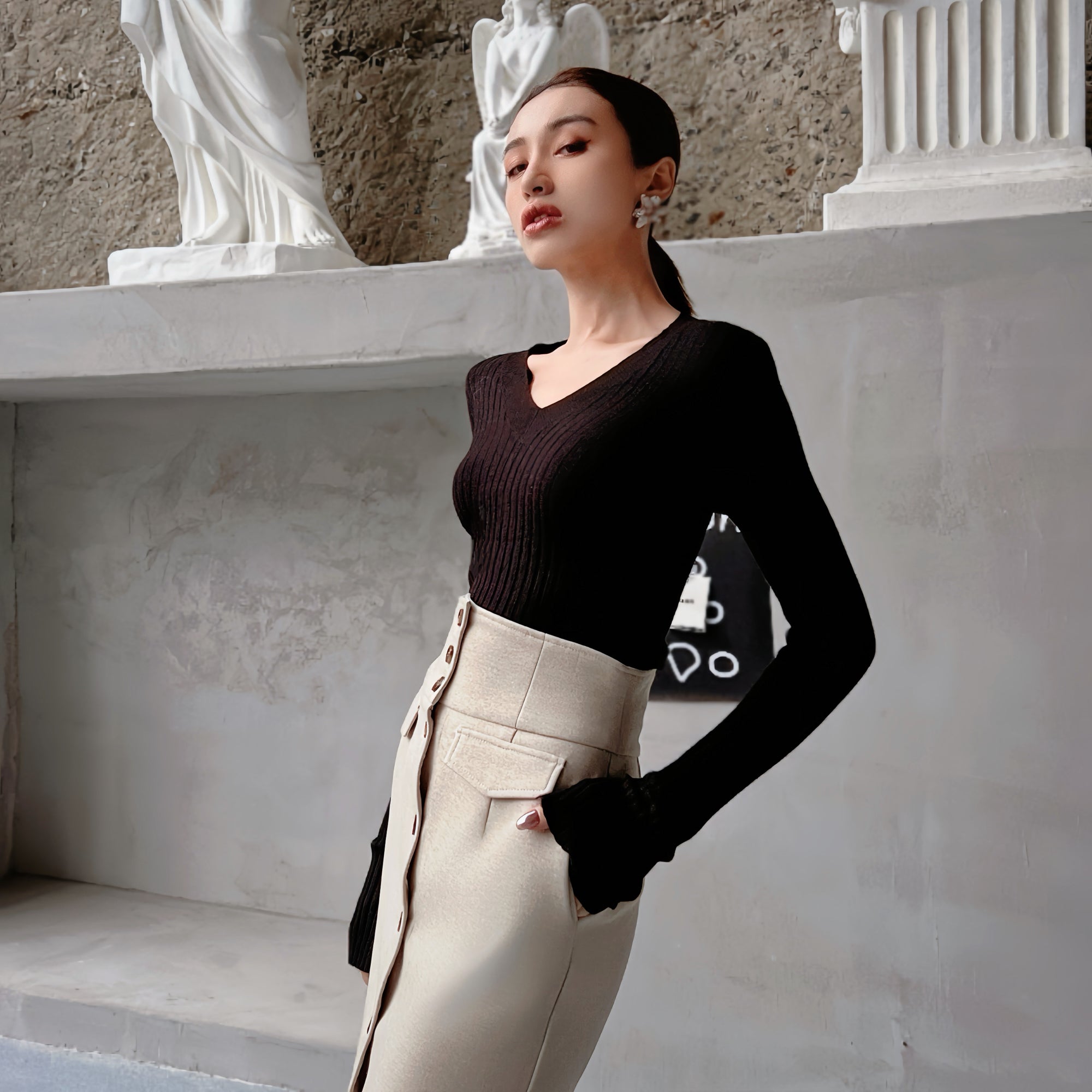 A woman wearing a black top and beige skirt stands in front of a white wall with statues.