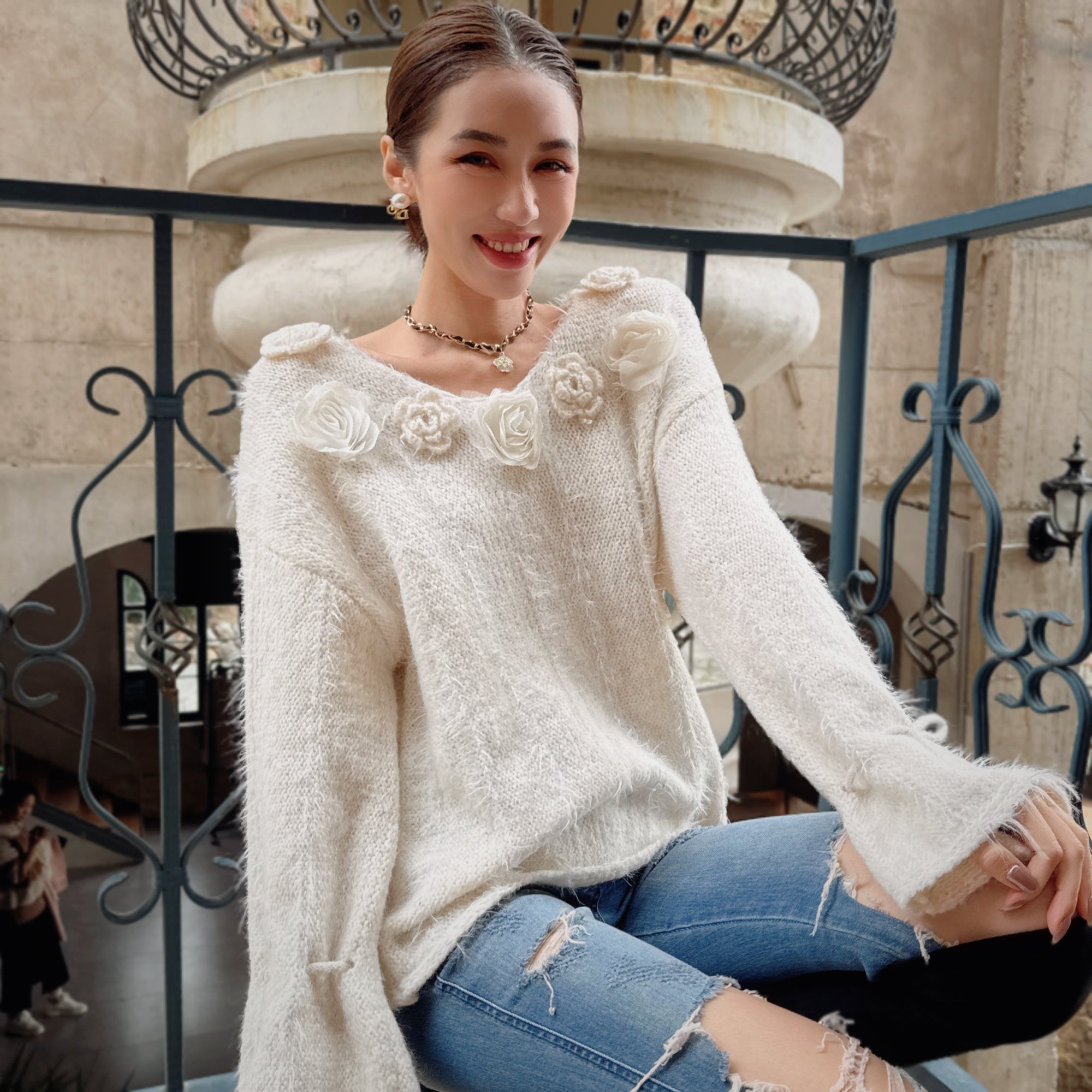 A woman wearing a white sweater with flower embellishments and distressed jeans is sitting on a railing, smiling and looking at the camera.