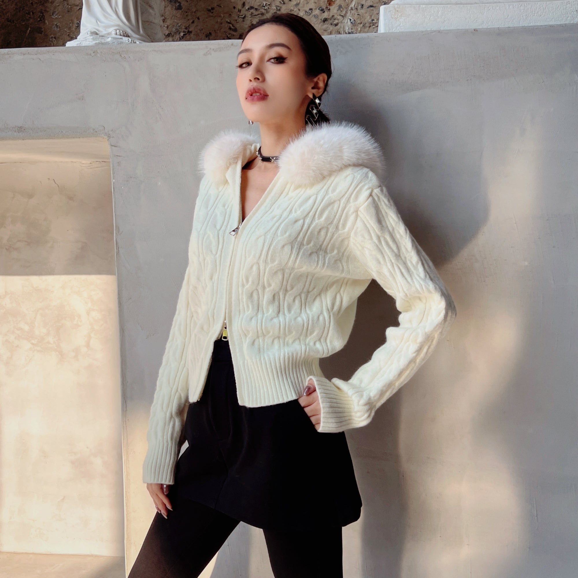 A woman wearing a white knitted cardigan with a fur collar and black skirt stands against a white wall, looking directly at the camera with a serious expression.