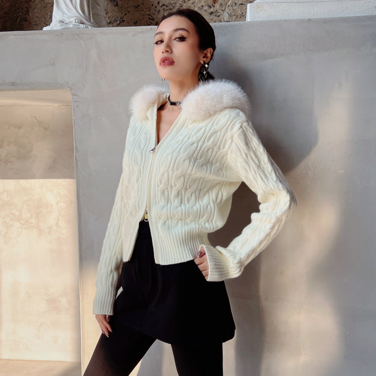A woman wearing a white knitted cardigan with a fur collar and black skirt stands against a white wall, looking directly at the camera with a serious expression.