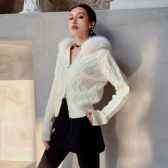 A woman wearing a white knitted cardigan with a fur collar and black skirt stands against a white wall, looking directly at the camera with a serious expression.