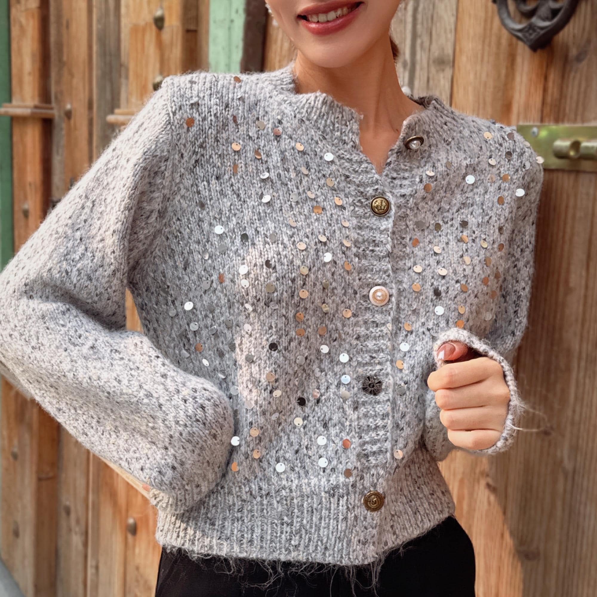 A woman wearing a gray sweater with silver buttons and a sparkly pattern stands in front of a wooden wall.