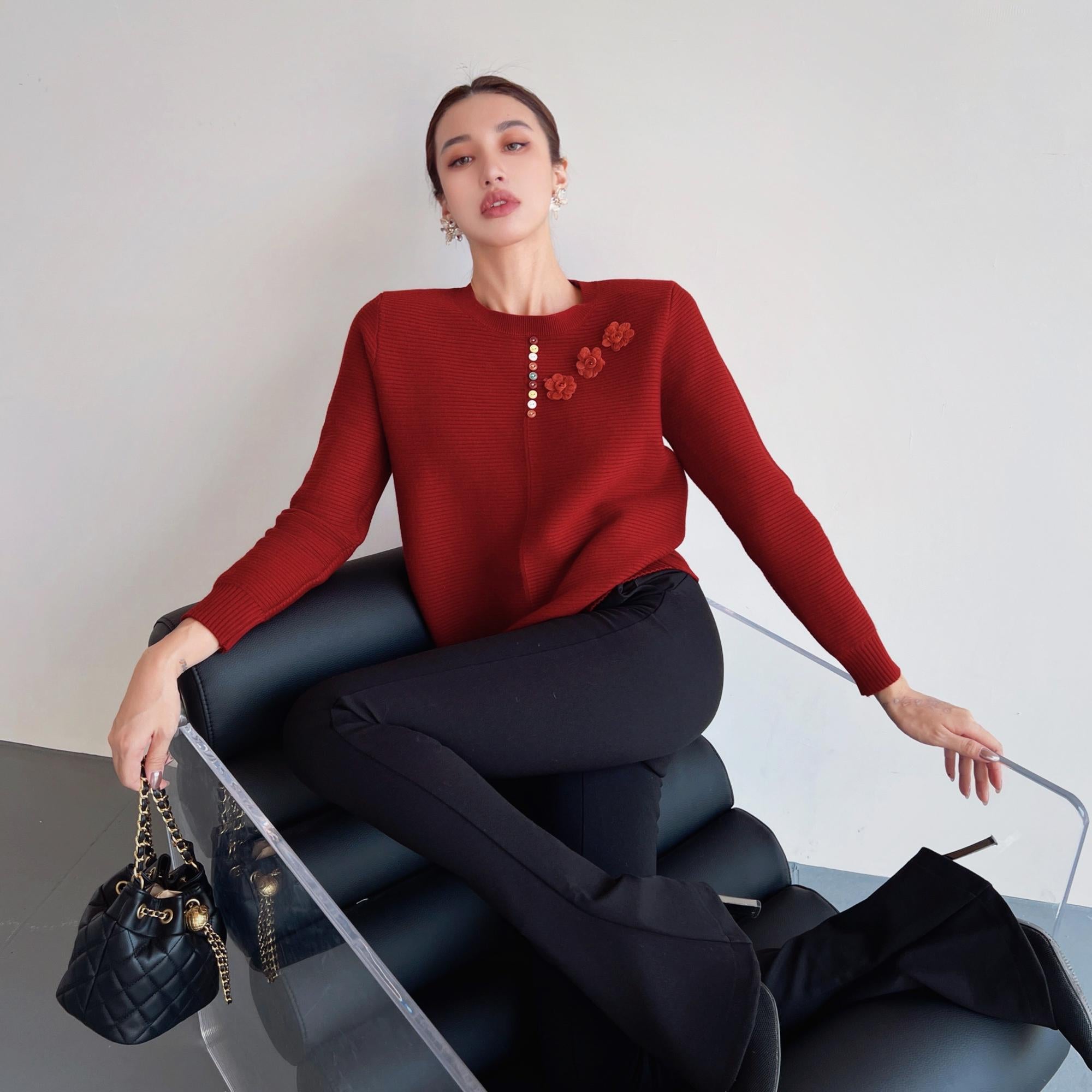 A woman wearing a red sweater with flower designs is sitting on a black chair, holding a black purse.