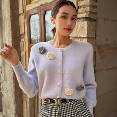 A woman wearing a light purple cardigan with white flower embellishments stands in front of a stone wall.