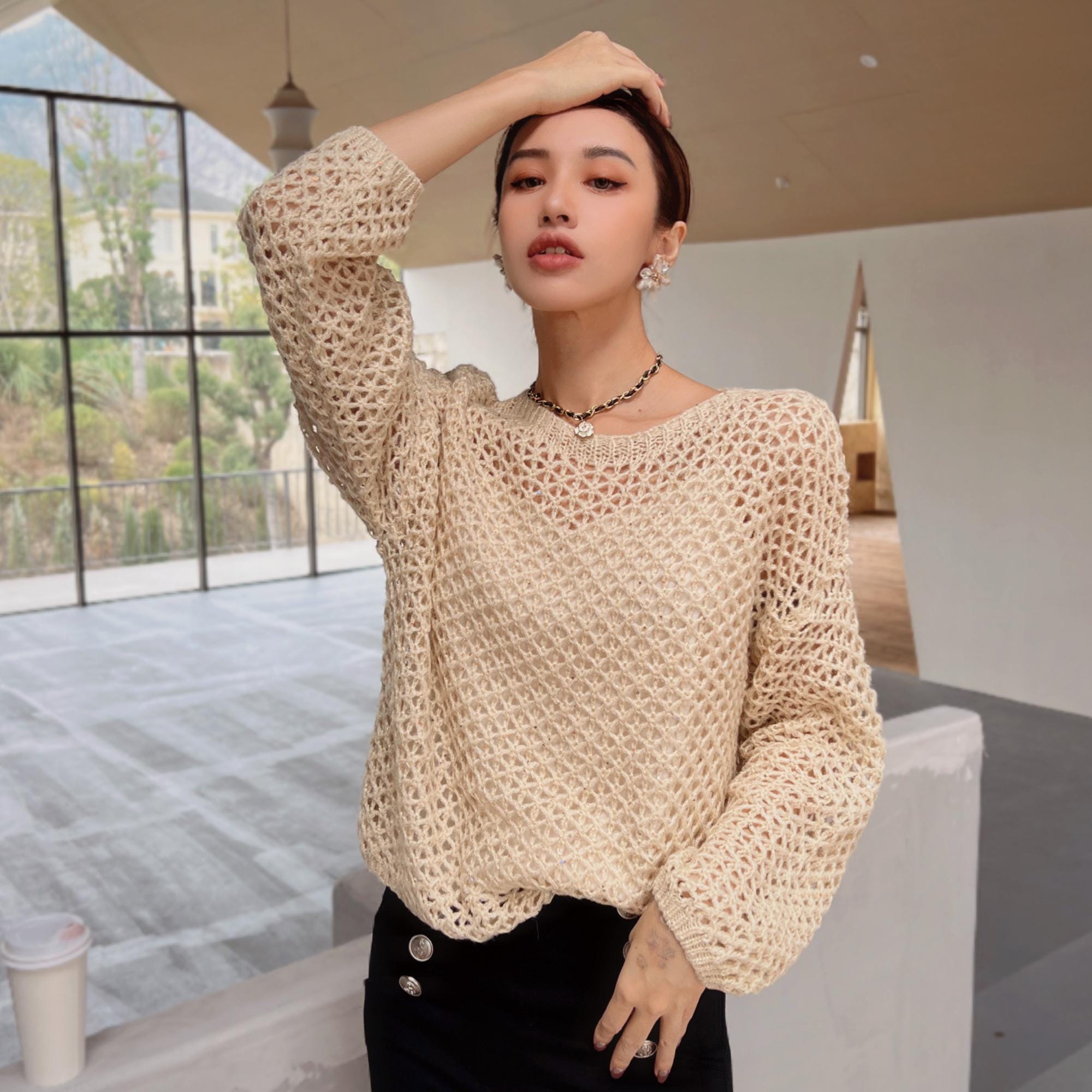 A woman wearing a beige crocheted sweater and black pants stands in a room with large windows, holding her hand up to her head.