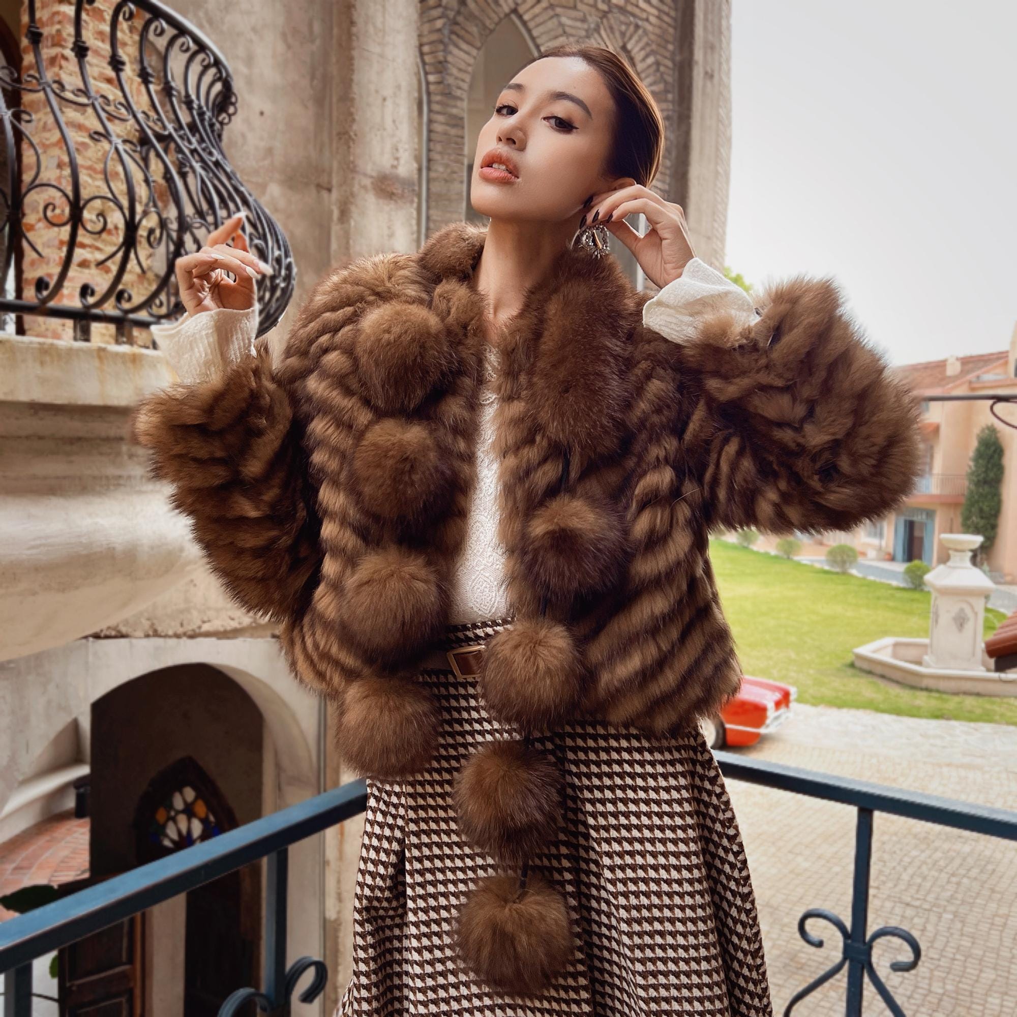 A woman wearing a brown fur coat and a patterned skirt stands on a balcony, with a stone building and a fountain visible in the background.