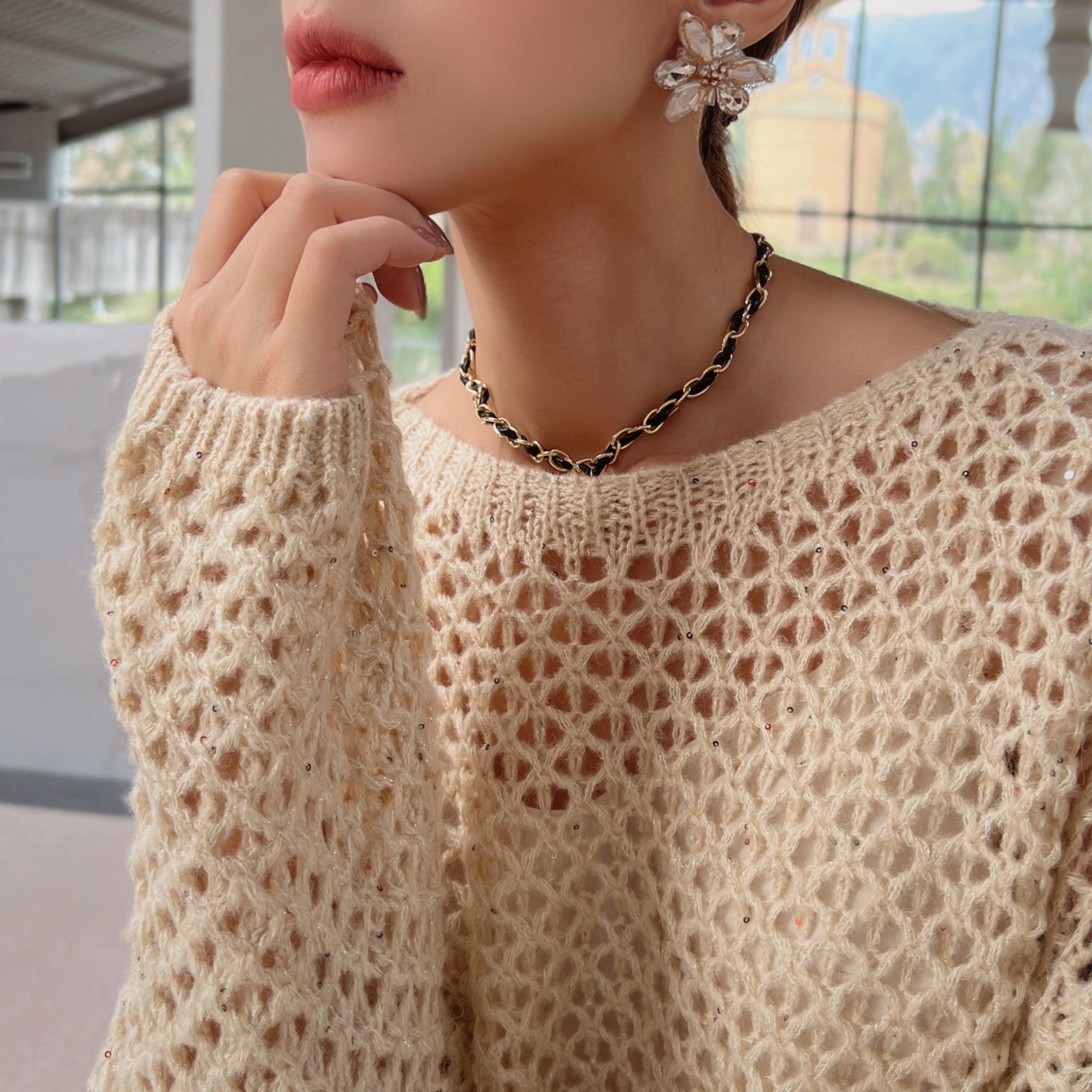 A close-up of a woman wearing a white lace sweater and a black and gold necklace, with her hand resting on her chin.