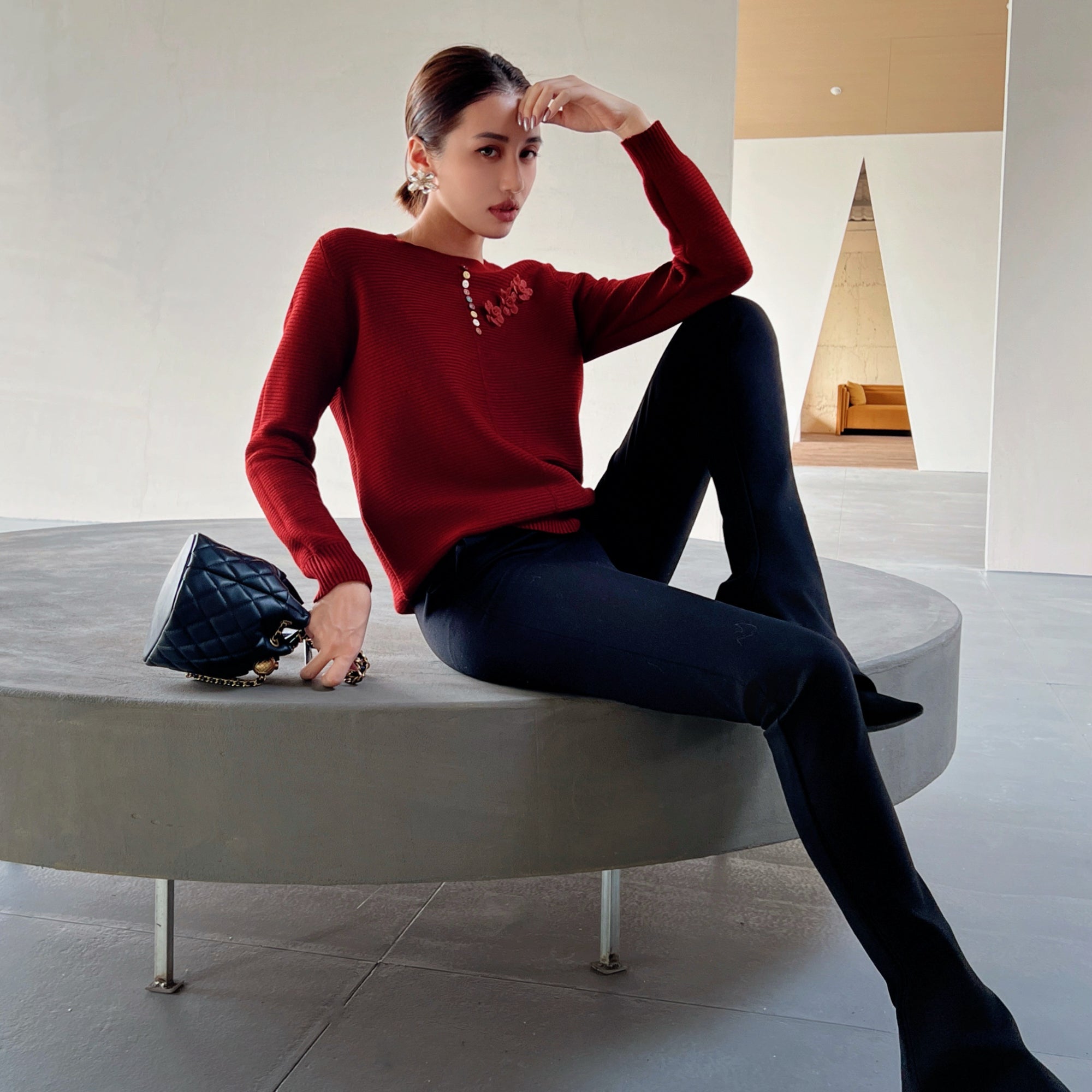 A woman in a red sweater and black pants sits on a circular gray bench, holding a black purse and looking directly at the camera.