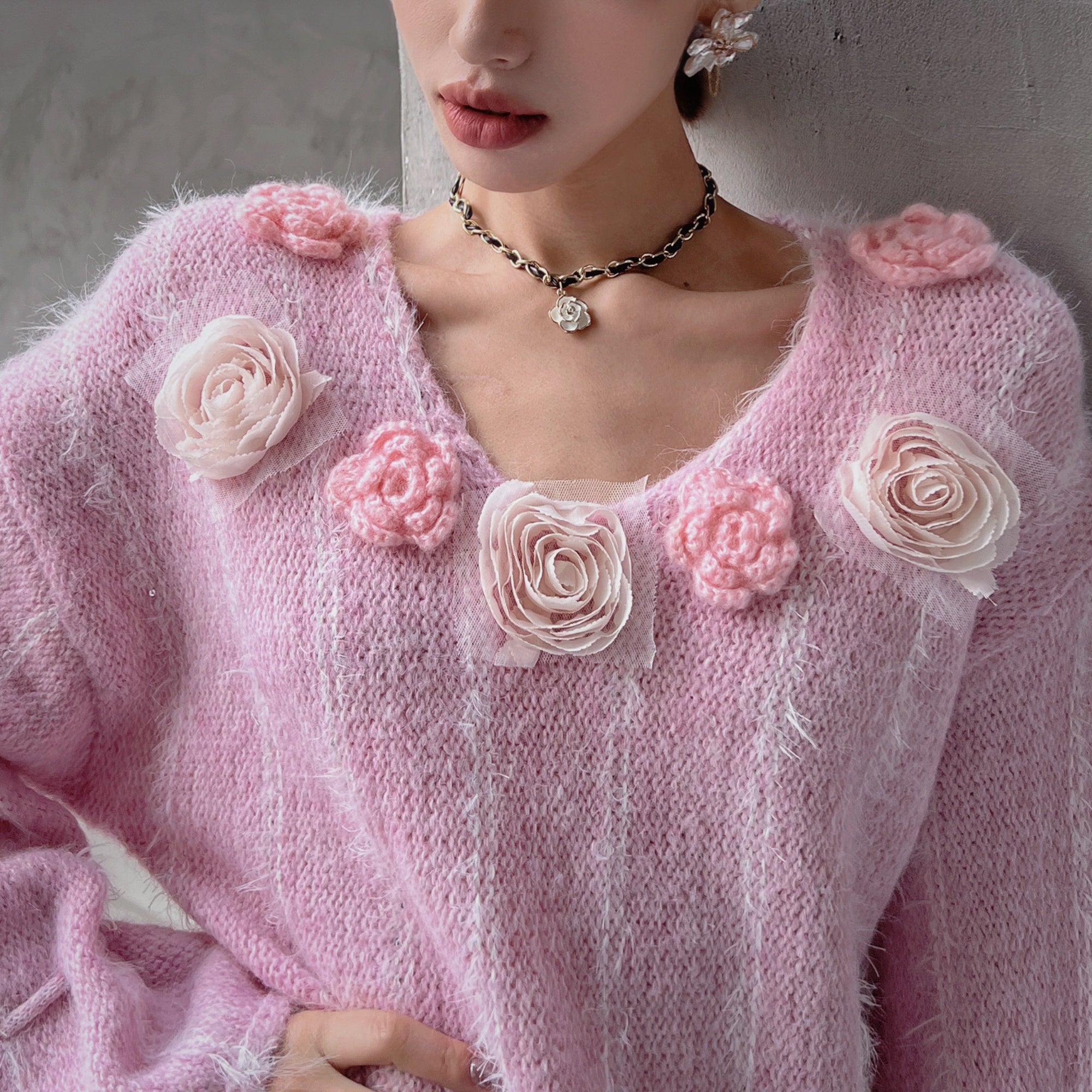 A woman wearing a pink sweater with white flowers on it, posing against a gray background.