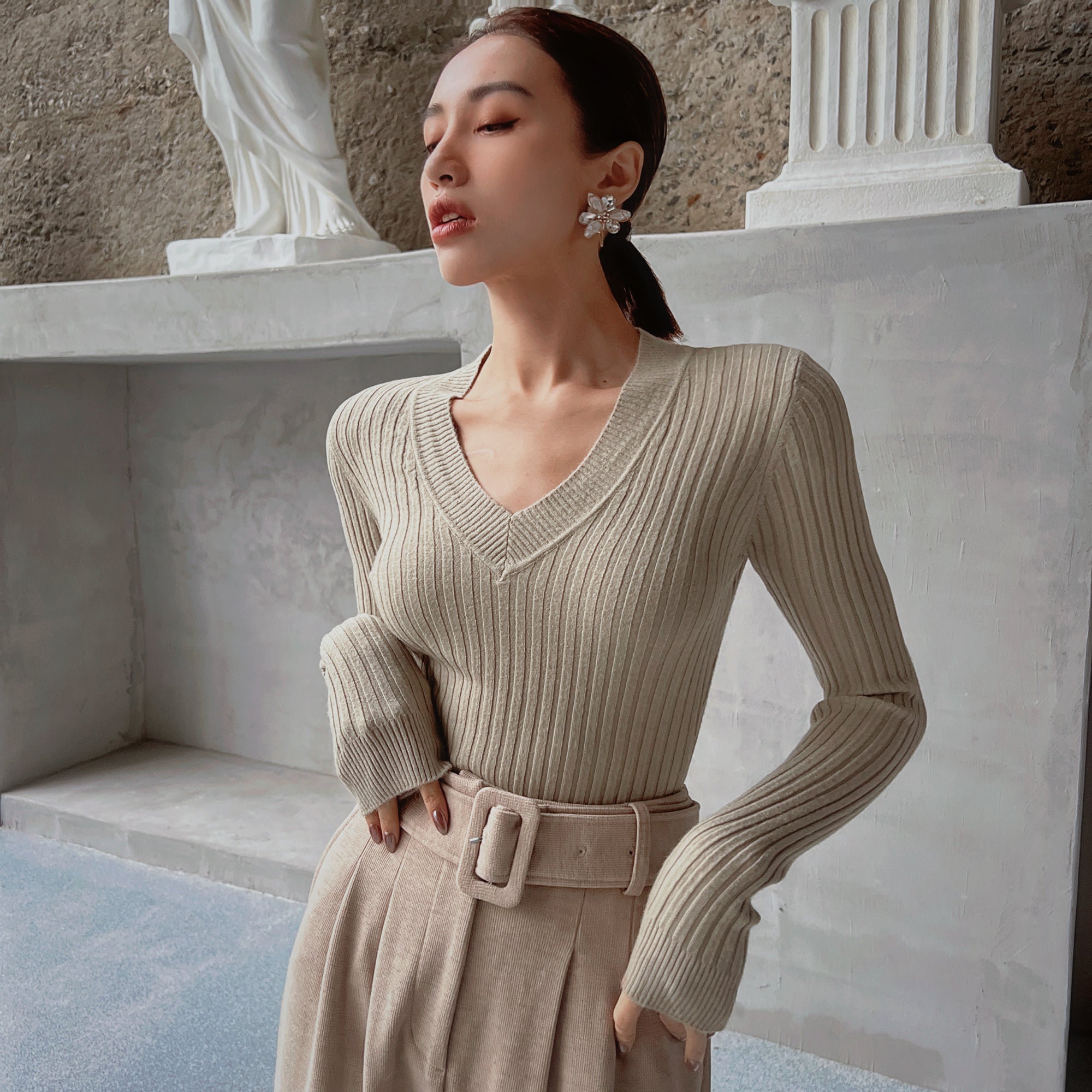 A woman wearing a beige sweater and a belt, with her eyes closed and looking away from the camera, standing in front of a white wall with a statue.