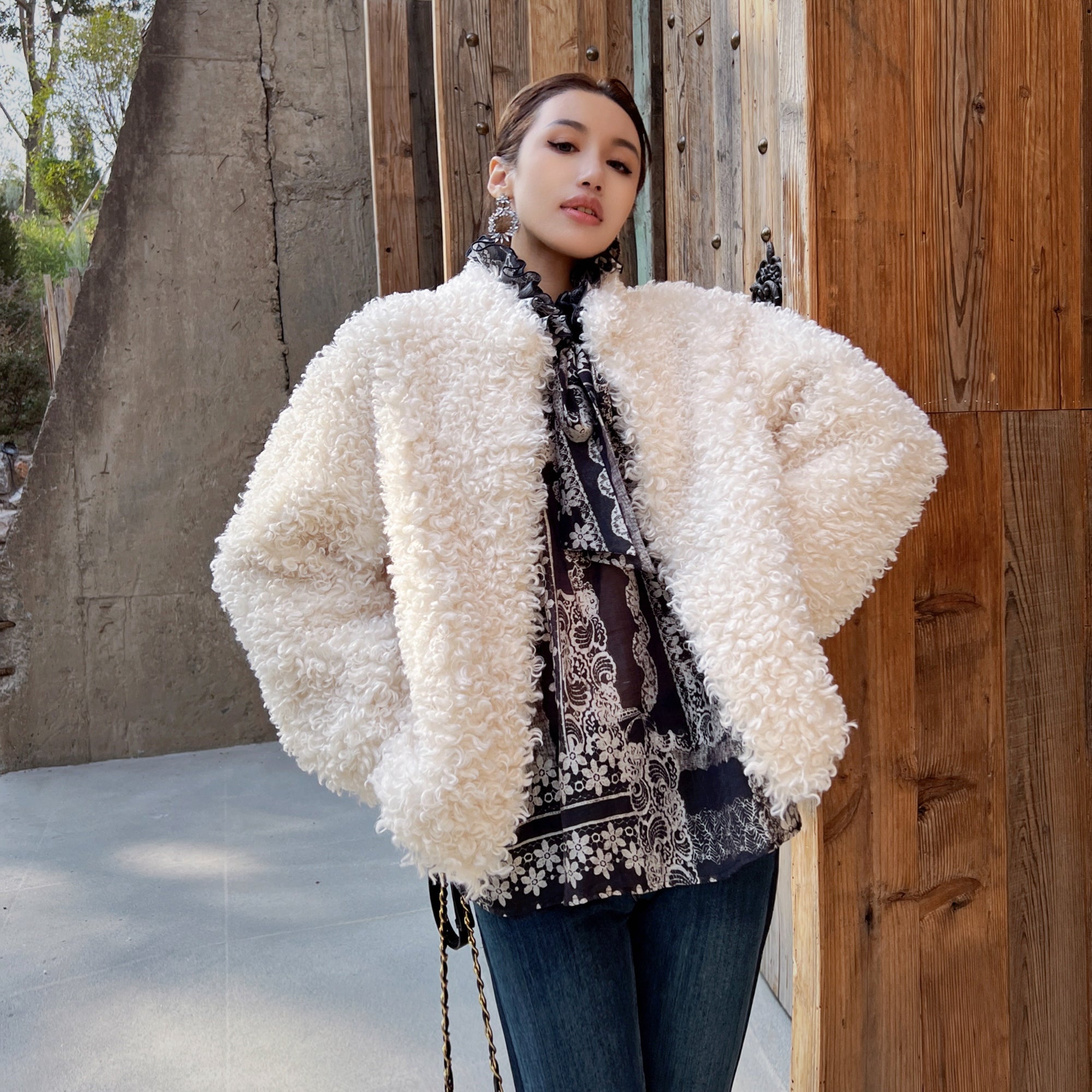 A woman wearing a white fluffy coat and a patterned shirt stands in front of a wooden wall.