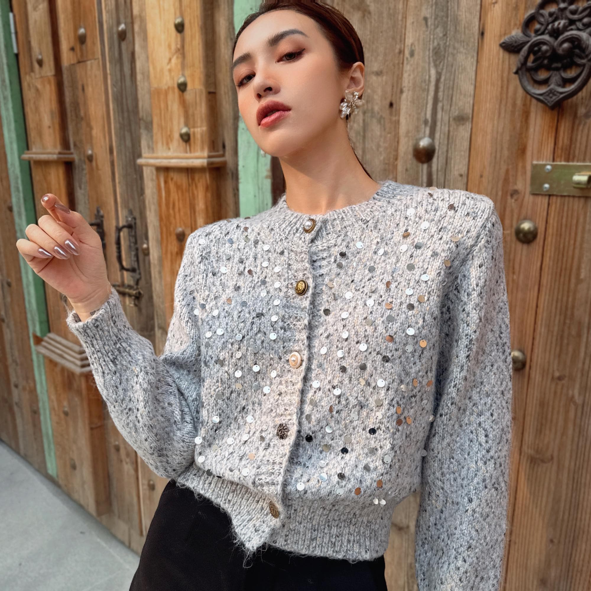 A woman wearing a grey sweater with silver buttons and a floral pattern stands in front of a wooden door.