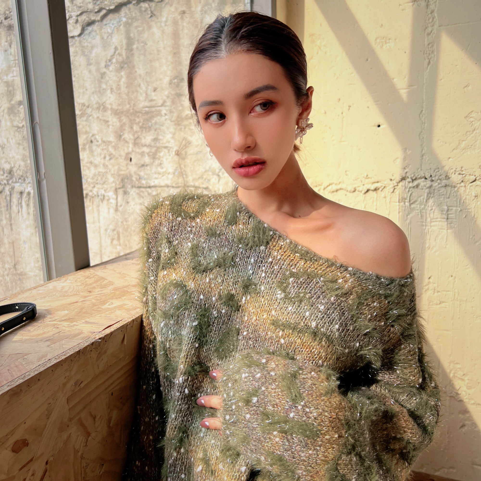 A woman wearing a green and gold patterned sweater stands in front of a window, looking off to the side with a thoughtful expression.