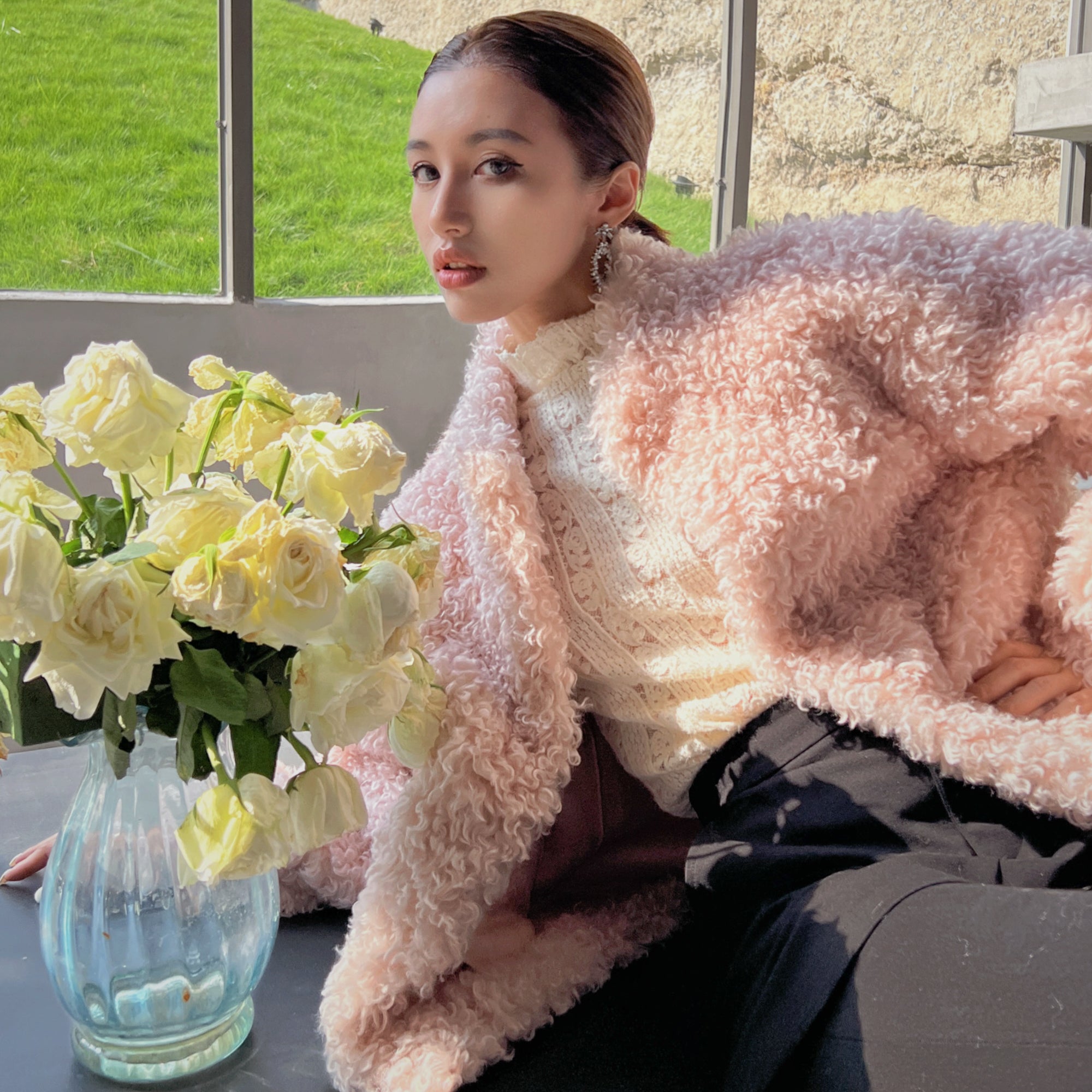 A woman in a pink coat sits next to a vase of white roses, with a window in the background.