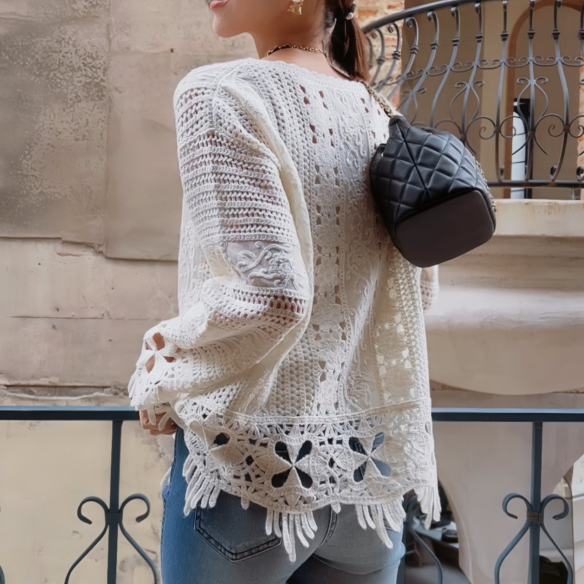 A woman wearing a white lace top with a floral pattern, blue jeans, and carrying a black quilted handbag stands in front of a building with a wrought iron railing.