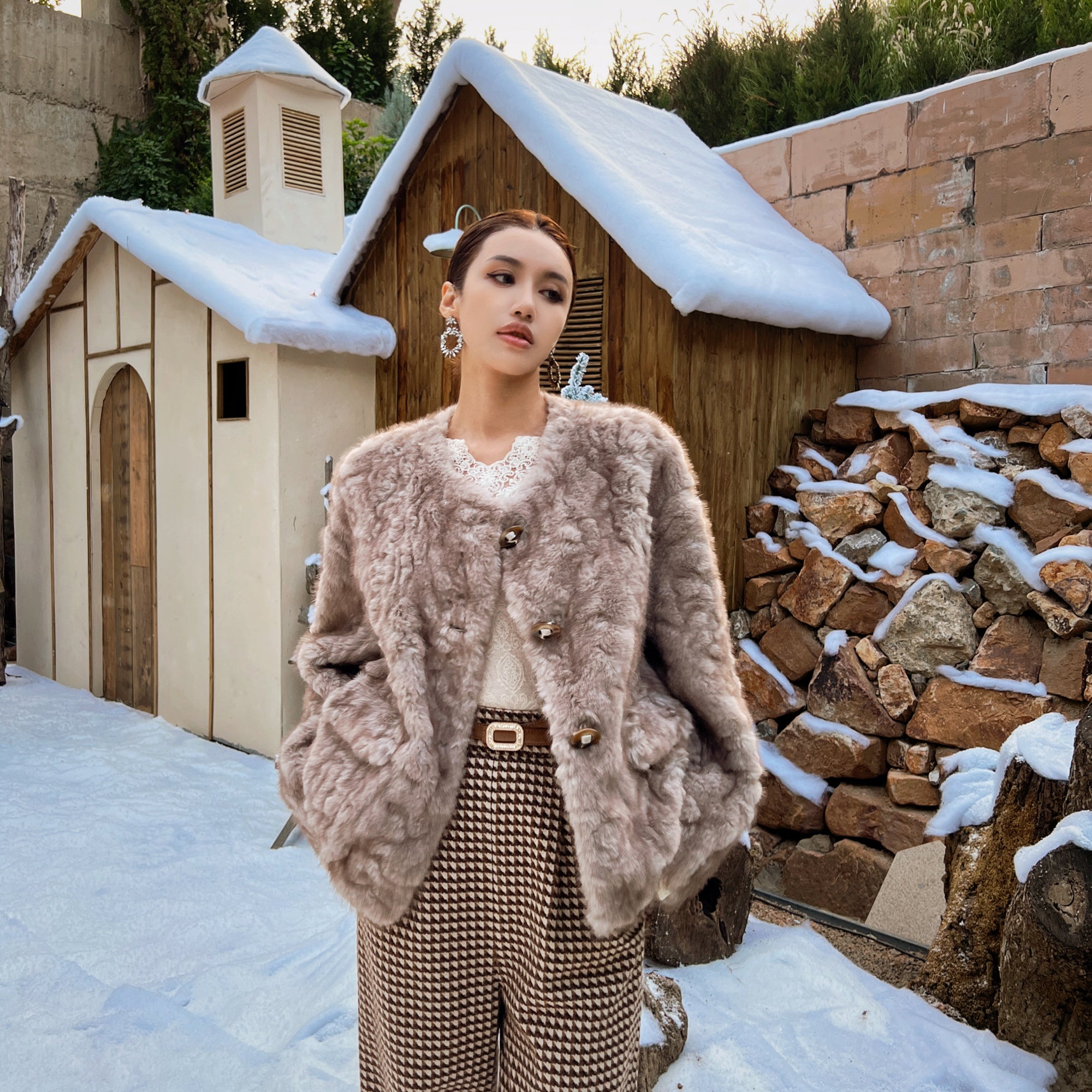 A woman wearing a fur coat and checkered pants stands in front of a snow-covered wooden structure, with a stone wall and trees in the background.