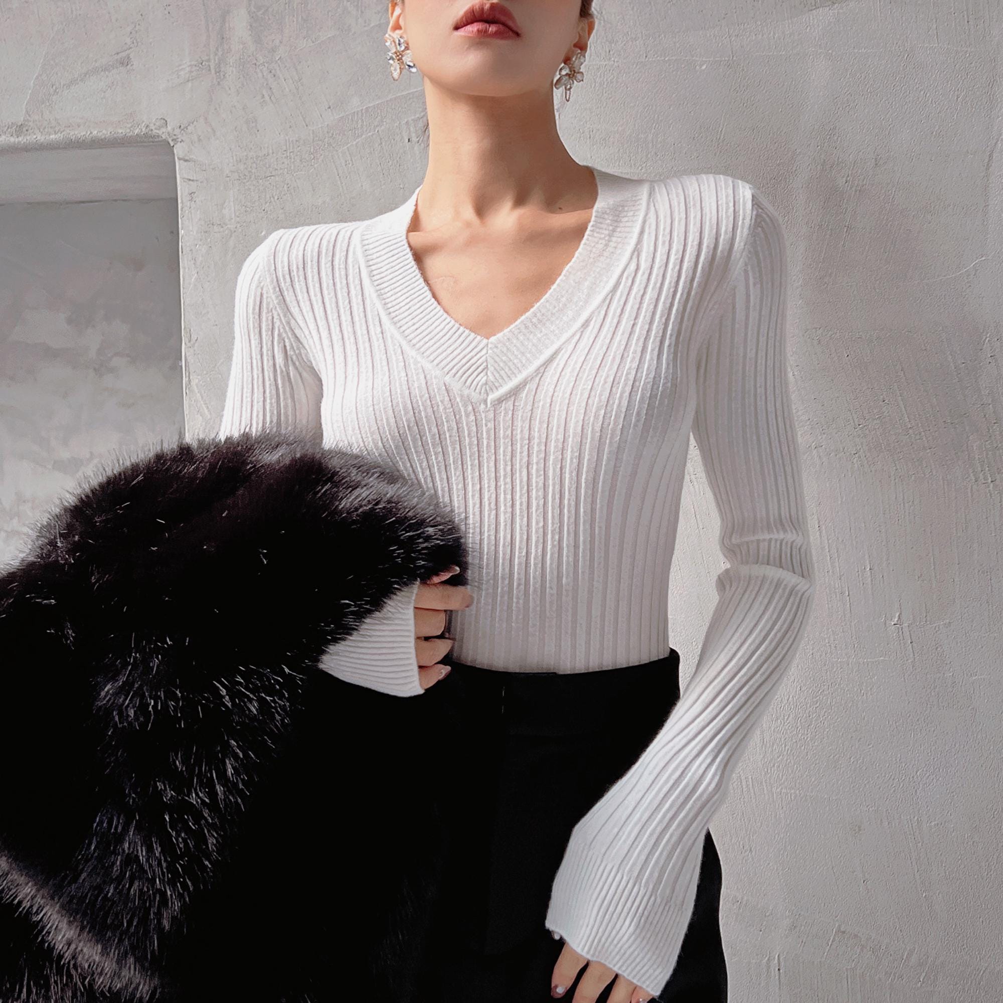 A woman wearing a white sweater and black pants stands against a white wall, holding a black fur coat.