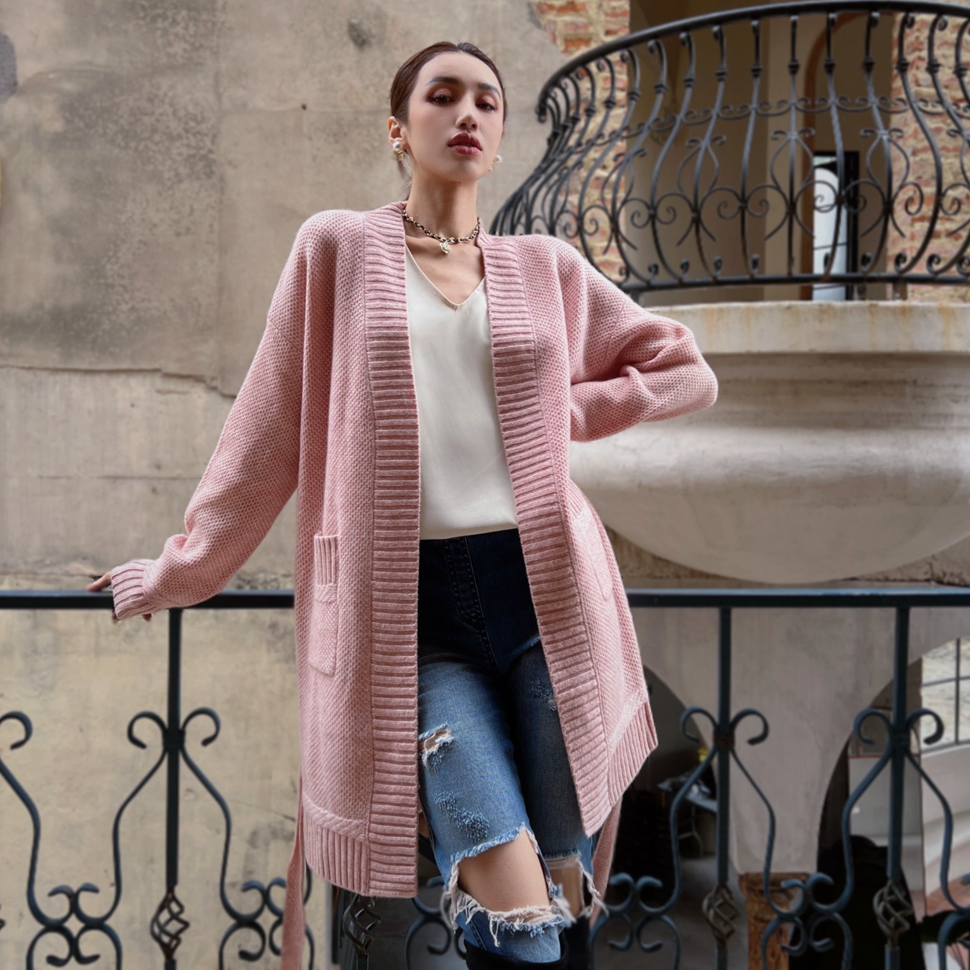 A woman wearing a pink cardigan and ripped jeans stands on a balcony, leaning against a wrought iron railing.