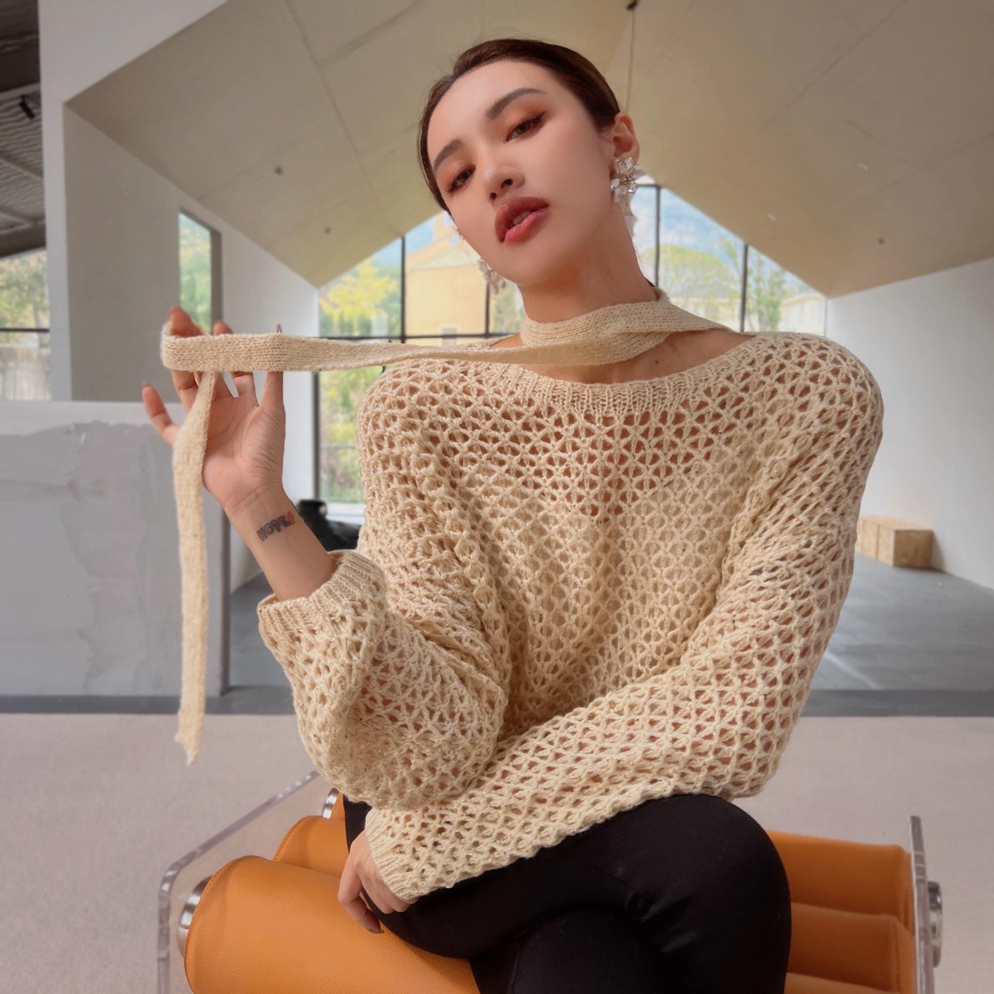 A woman wearing a beige sweater and holding a scarf around her neck is sitting on an orange chair in a room with large windows.