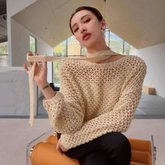A woman wearing a beige sweater and holding a scarf around her neck is sitting on an orange chair in a room with large windows.