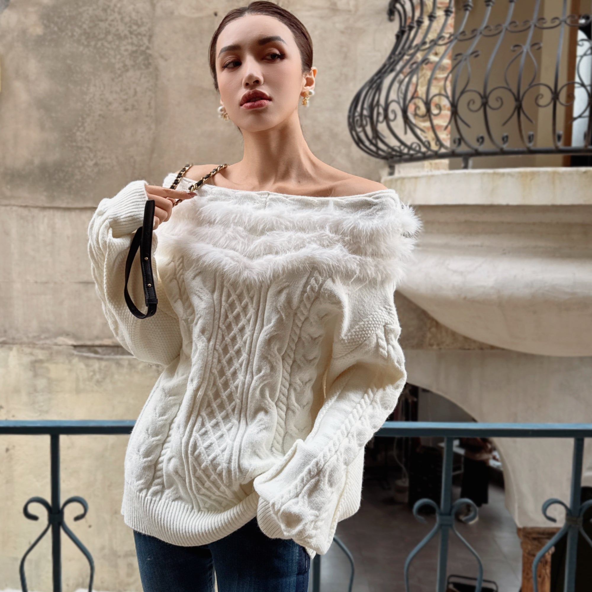 A woman wearing a white knitted sweater with a fur collar stands in front of a wrought iron balcony.