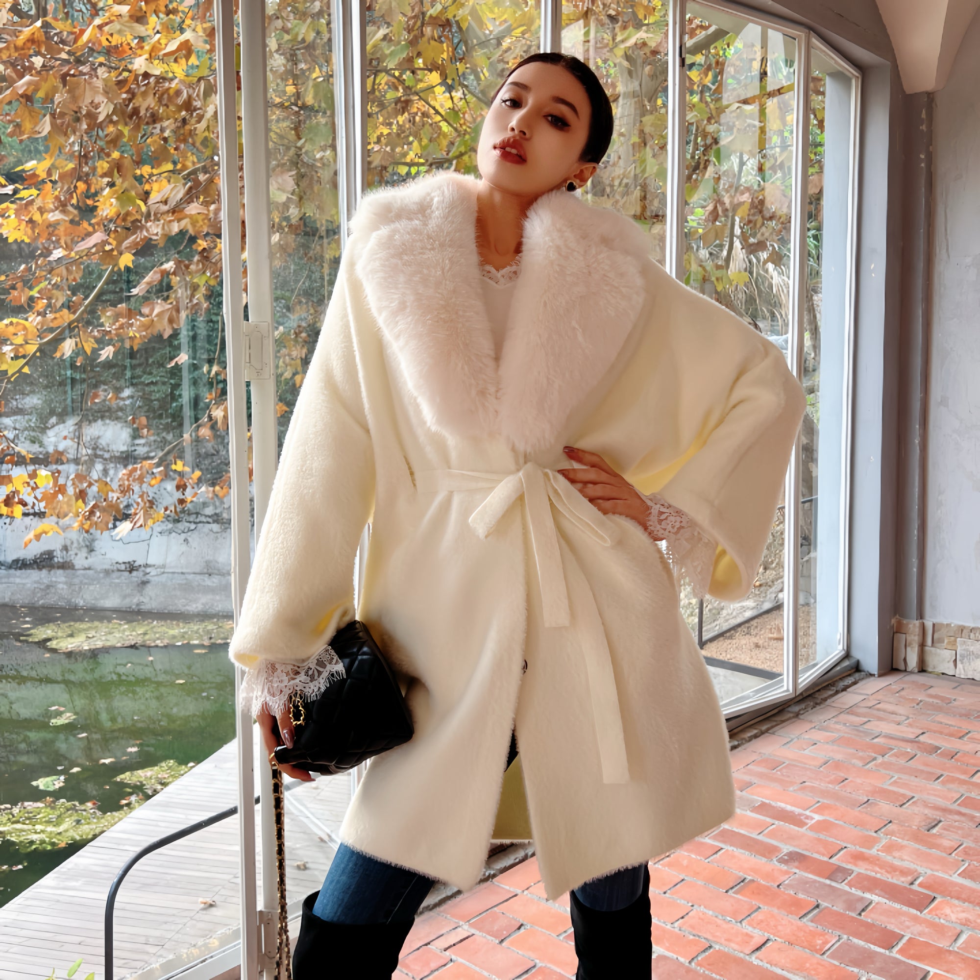 A woman wearing a white fur coat with a fur collar and holding a black purse stands in front of a window with autumn trees visible outside.