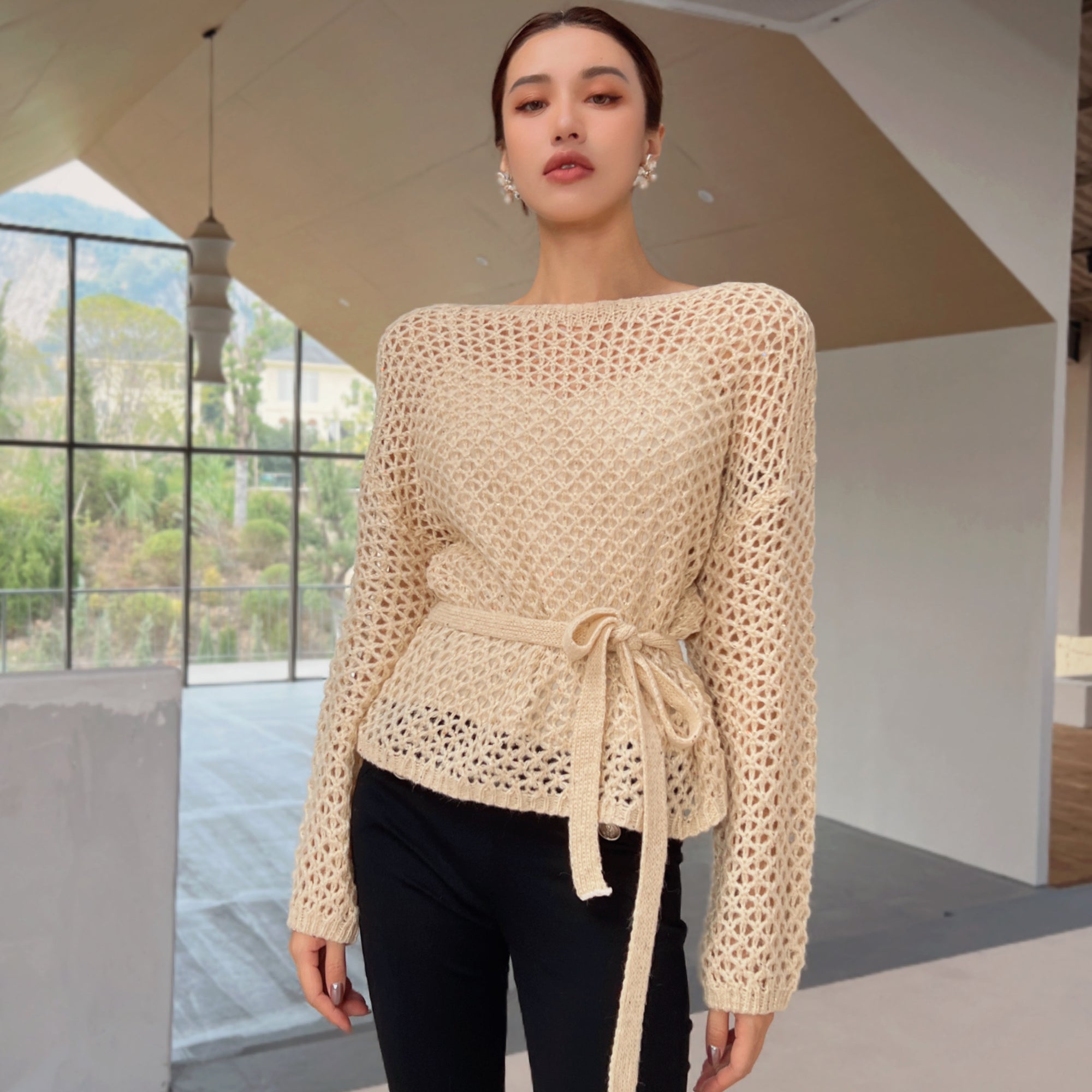 A woman wearing a beige crocheted sweater with a tie, standing in an indoor setting with a large window and a hanging light fixture.
